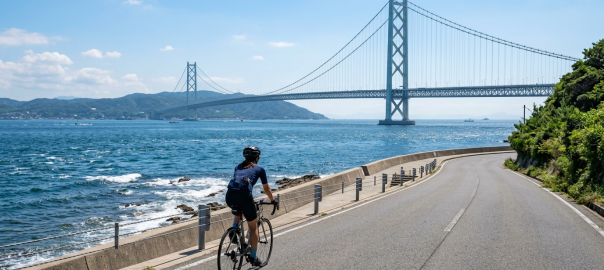 單車騎士沿淡路島海岸道路騎乘，遠方可見明石海峽大橋（AI 生成示意圖）