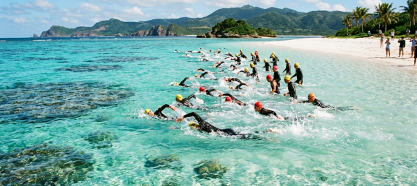 鐵人三項選手於沖繩久米島清澈見底的珊瑚礁白砂海域破浪出發海泳（AI 生成示意圖）